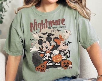 May include: Sage green t-shirt featuring a Halloween-themed graphic with Mickey and Minnie Mouse in skeleton costumes, pumpkins, ghosts, and bats. The text "Nightmare on Main Street" is printed above the characters. The shirt is a relaxed fit.