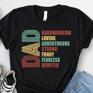 May include: Black t-shirt with a retro-style graphic that says "DAD" with the letters stacked vertically. The graphic is made up of colorful stripes and the words "Hardworking, Loving, Adventurous, Strong, Funny, Fearless, Devoted" are listed below the letters.