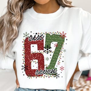 Może przedstawiać: Biała bluza z nadrukiem świątecznym. Grafika przedstawia liczby "67" w kolorze czerwonym i zielonym, ze słowami "six" i "seven" w skrypcie. Projekt jest ozdobiony płatkami śniegu i lampkami.
