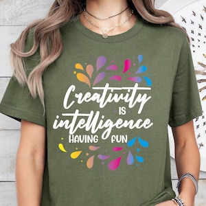 May include: A green t-shirt with a white graphic that says "Creativity is intelligence having fun". The graphic is surrounded by colorful water drop shapes.