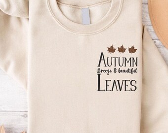 May include: A cream-colored sweatshirt with the text "Autumn Breeze & Beautiful Leaves" and three brown maple leaf graphics. The sweatshirt is folded, with a tan hat visible in the background.