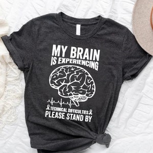 May include: A dark gray t-shirt with a white graphic of a brain with a heartbeat line and the text "My brain is experiencing technical difficulties. Please stand by."