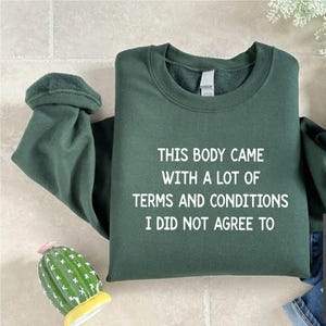May include: A dark green sweatshirt with the text "THIS BODY CAME WITH A LOT OF TERMS AND CONDITIONS I DID NOT AGREE TO" printed in white. The sweatshirt is folded and laid flat on a light brown surface.