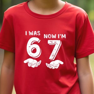 May include: A red t-shirt with the text "I WAS 6 NOW I'M 67" in white. The shirt features the numbers 6 and 7 with a hand graphic underneath each number. The person wearing the shirt is smiling.