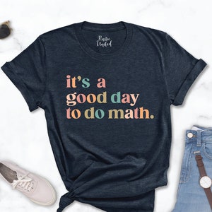 May include: A navy blue t-shirt with the text "it's a good day to do math." in rainbow colors. The shirt is laid flat on a white marble surface with a pair of sunglasses, two white sneakers, and a pair of blue jeans with a brown watch.