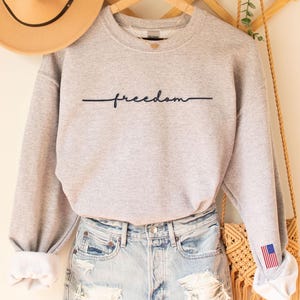 May include: A light gray sweatshirt with the word "freedom" in black script. The sweatshirt is paired with denim shorts. A tan hat and a macrame bag with an American flag detail are also visible.
