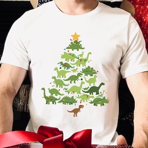 May include: White t-shirt featuring a Christmas tree design made of green dinosaurs, with a yellow star on top. The shirt is worn by a person holding a red gift box with a large red bow.