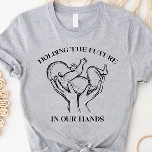 May include: Gray t-shirt with a black and white illustration of a baby in hands. The text "Holding the future in our hands" is printed on the shirt.