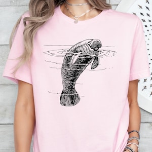 May include: A pink t-shirt with a black and white illustration of a manatee swimming in the water.