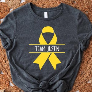May include: Dark gray t-shirt featuring a yellow awareness ribbon design with the text "TEAM JUSTIN". The shirt is made of a soft material and has a crew neck. The shirt is displayed on a brown woven surface.