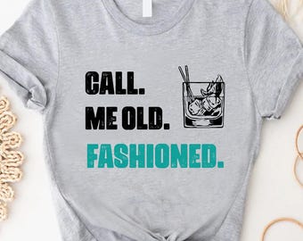 May include: A light gray t-shirt with the text "CALL ME OLD. FASHIONED." in black and teal. A graphic of a cocktail glass with ice and a straw is printed on the shirt. The shirt is knotted at the bottom.