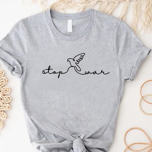 May include: A light gray t-shirt featuring a black line drawing of a dove with the words "stop war" in a handwritten font. The shirt is knotted at the waist.