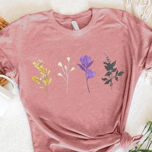 May include: A pink t-shirt with a floral design featuring four different flowers in yellow, white, purple, and black.