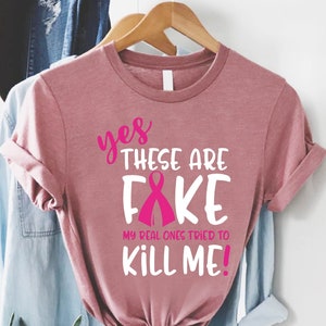 May include: Pink t-shirt with the text "yes these are FAKE my real ones tried to KILL ME!" with a pink ribbon graphic.