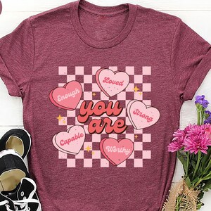May include: Heather maroon t-shirt with a graphic design featuring the words "You Are" in a retro font, surrounded by pink heart-shaped candies with positive affirmations. Black sneakers and a bouquet of flowers are also in the image.