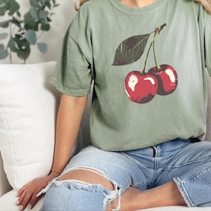 May include: Sage green t-shirt featuring a graphic of two red cherries with a green leaf. The shirt is worn with blue jeans. The overall theme is casual wear.