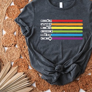 Puede incluir: Una camiseta gris oscuro con un dise&ntilde;o de rayas arco&iacute;ris que presenta una serie de siluetas de sables de luz blancas.