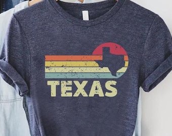 May include: Navy blue t-shirt with a retro design featuring the state of Texas. The design includes a red sun, horizontal stripes in orange, yellow, and teal, and the word "TEXAS" in a vintage font. The shirt is hanging on a wooden hanger.