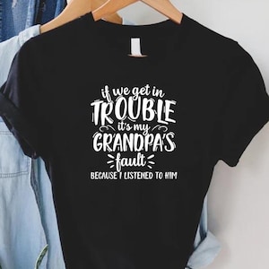 May include: Black t-shirt with the white text "if we get in trouble it's my grandpa's fault because I listened to him." The shirt is hanging on a wooden hanger with a denim shirt and a brown hat in the background.
