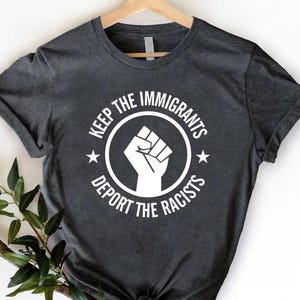 May include: Dark gray t-shirt with a white graphic. The graphic features a clenched fist inside a circle with the text "KEEP THE IMMIGRANTS" above and "DEPORT THE RACISTS" below. Two stars flank the fist.