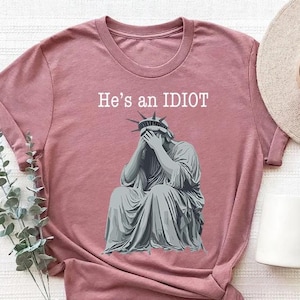 May include: Dusty pink t-shirt with a distressed graphic of the Statue of Liberty covering her face, and the text "He's an IDIOT" in white. A hat and mug are visible in the background. The shirt is folded.