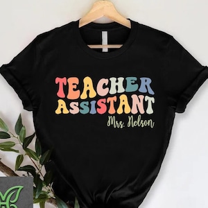 May include: Black t-shirt with the text "Teacher Assistant" in rainbow colors and the name "Mrs. Nelson" in black below.