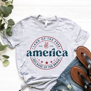 May include: A gray t-shirt with a red, white, and blue graphic design that says "Land of the Free, Since 1776, America, Because of the Brave". The graphic features a peace sign and stars.