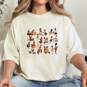 May include: A white t-shirt with a graphic print of a diverse group of people in various poses. The graphic features people of different skin tones and body types, all holding hands and smiling.