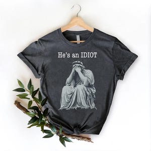 May include: Dark grey t-shirt featuring the text "He's an IDIOT" above a grey illustration of the Statue of Liberty with her face in her hands. The t-shirt is displayed on a wooden hanger.