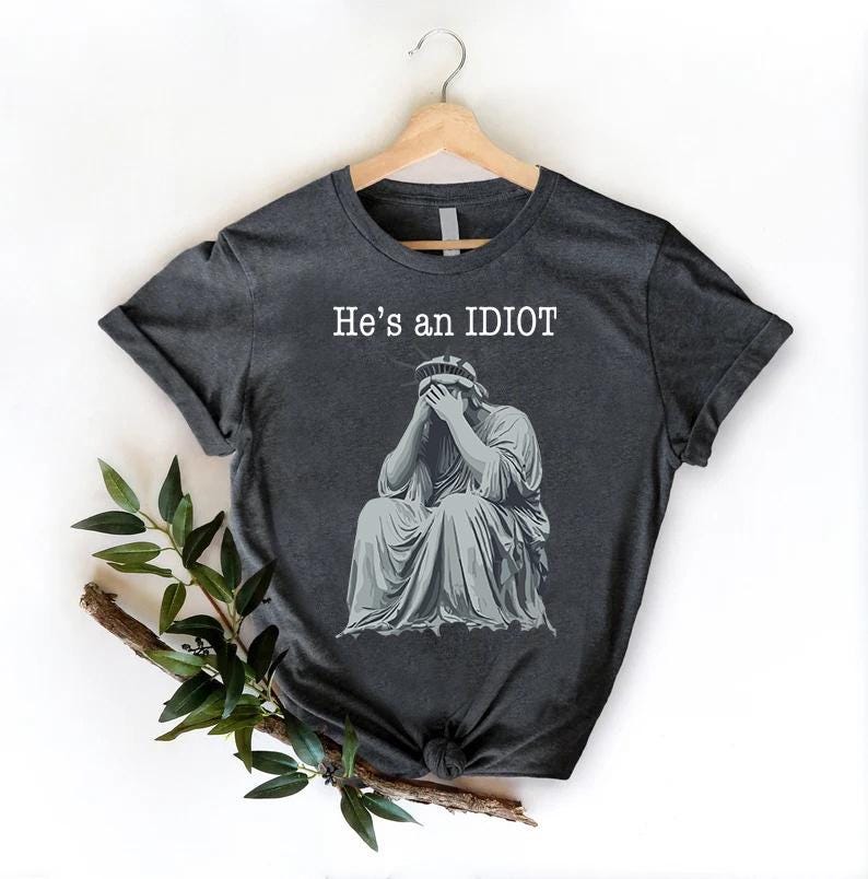 May include: Dark grey t-shirt featuring the text "He's an IDIOT" above a grey illustration of the Statue of Liberty with her face in her hands. The t-shirt is displayed on a wooden hanger.