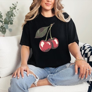 May include: Black t-shirt featuring a graphic of two red cherries with a green leaf. The shirt is worn with blue jeans. The cherries have a vintage, slightly distressed look.