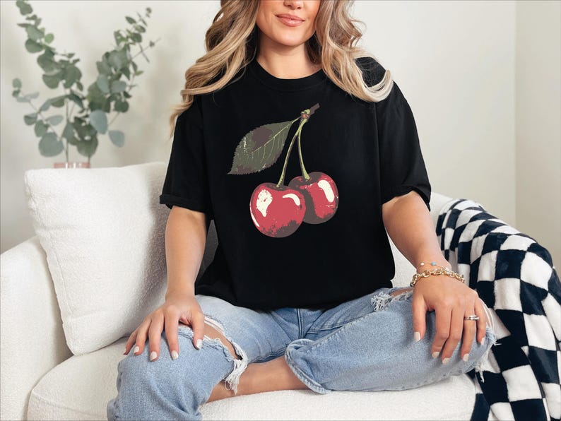 May include: Black t-shirt featuring a graphic of two red cherries with a green leaf. The shirt is worn with blue jeans. The cherries have a vintage, slightly distressed look.