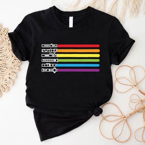 Puede incluir: Camiseta negra con un dise&ntilde;o de arco&iacute;ris de sables de luz. Los colores del arco&iacute;ris son rojo, naranja, amarillo, verde, azul, a&ntilde;il y violeta.
