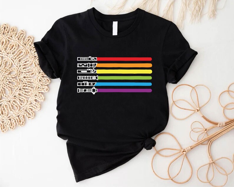 Puede incluir: Camiseta negra con un dise&ntilde;o de arco&iacute;ris de sables de luz. Los colores del arco&iacute;ris son rojo, naranja, amarillo, verde, azul, a&ntilde;il y violeta.