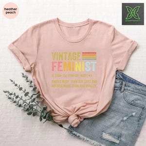 May include: A pink t-shirt with the text "Vintage Feminist" in rainbow colors. The text "N. (VIN-TIJ FEM-UH-NIST)" is below the main text, followed by the text "Knows more than she says and notices more than you realize".