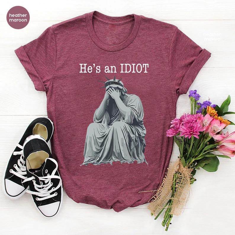 May include: Heather maroon t-shirt with the text "He's an IDIOT" above a grey illustration of the Statue of Liberty covering her face. Black and white trainers are on the left. A bouquet of flowers is on the right.