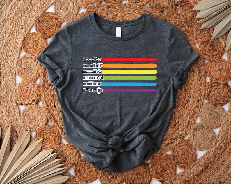 Puede incluir: Una camiseta gris oscuro con un dise&ntilde;o de rayas arco&iacute;ris que presenta una serie de siluetas de sables de luz blancas.