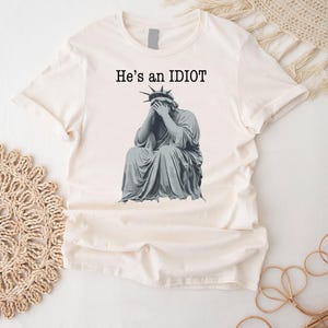 May include: A cream-coloured t-shirt with a graphic of the Statue of Liberty covering her face. The text "He's an IDIOT" is printed above the image in black. The shirt is made of a soft, lightweight material. A casual, everyday garment.