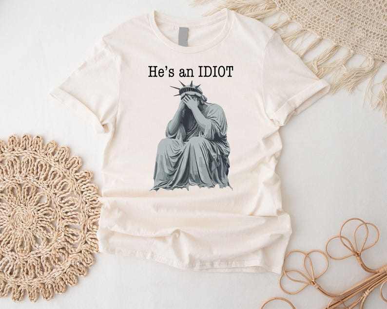 May include: A cream-coloured t-shirt with a graphic of the Statue of Liberty covering her face. The text "He's an IDIOT" is printed above the image in black. The shirt is made of a soft, lightweight material. A casual, everyday garment.