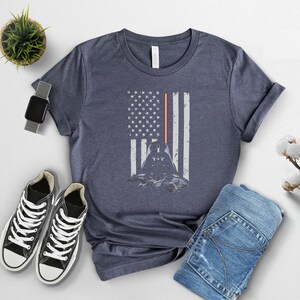 Puede incluir: Camiseta azul marino con un dise&ntilde;o de Darth Vader integrado en una bandera estadounidense. La bandera incluye un sable de luz rojo y una silueta del personaje. La camiseta se combina con zapatillas negras y vaqueros azules.