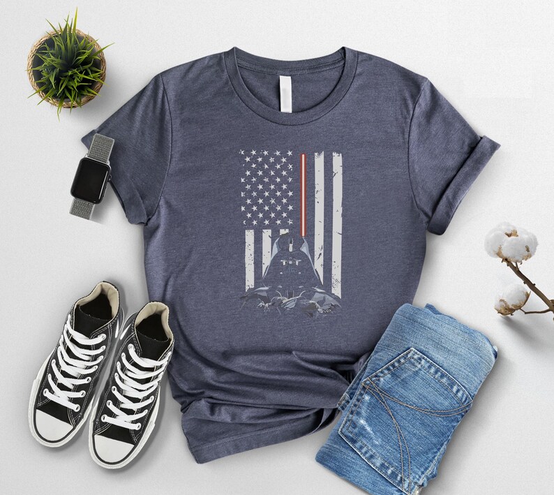 Puede incluir: Camiseta azul marino con un dise&ntilde;o de Darth Vader integrado en una bandera estadounidense. La bandera incluye un sable de luz rojo y una silueta del personaje. La camiseta se combina con zapatillas negras y vaqueros azules.
