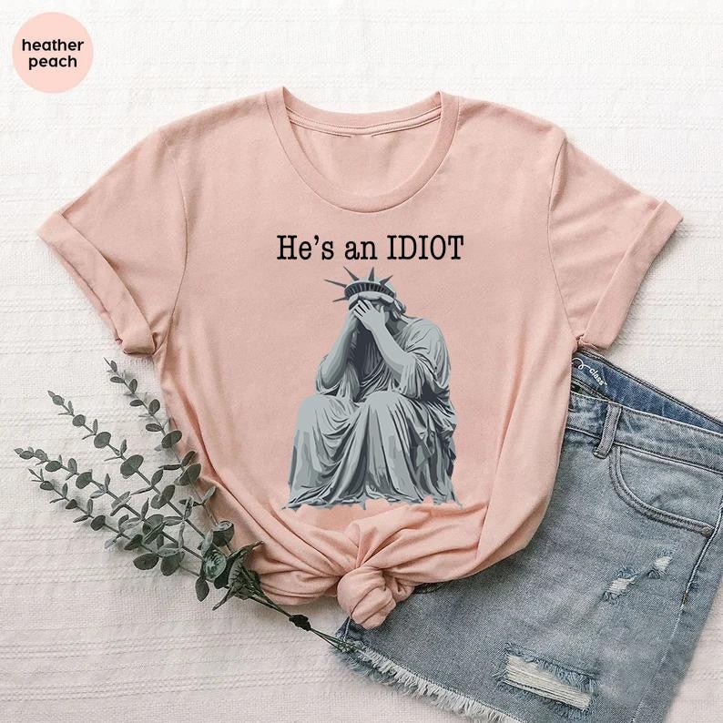 May include: A pale pink t-shirt with a graphic of the Statue of Liberty covering her face. The text "He's an IDIOT" is printed above the statue in black. The shirt is styled with denim shorts.