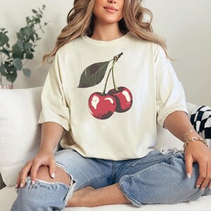 May include: Cream-colored t-shirt featuring a vintage-style illustration of two red cherries with a green leaf. The shirt is paired with ripped blue jeans. The overall aesthetic is casual and relaxed.