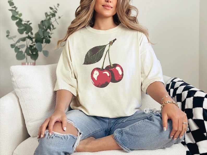 May include: Cream-colored t-shirt featuring a vintage-style illustration of two red cherries with a green leaf. The shirt is paired with ripped blue jeans. The overall aesthetic is casual and relaxed.