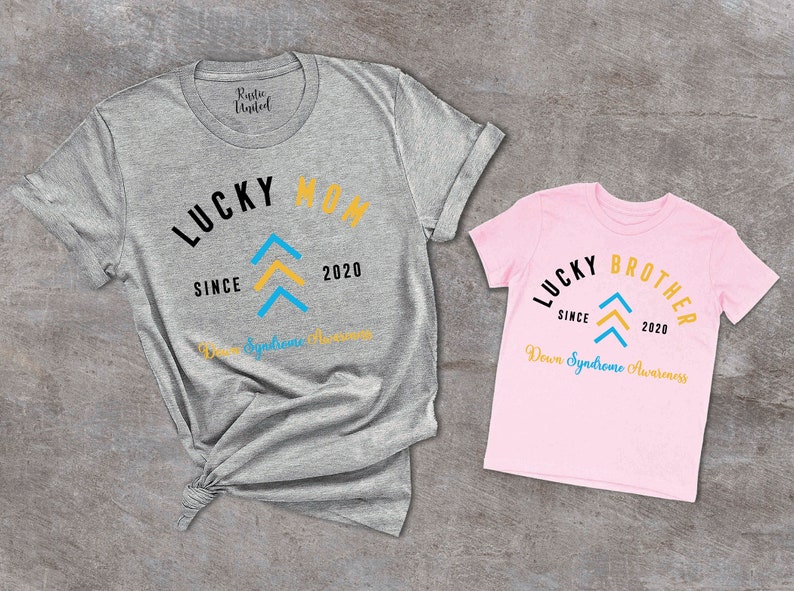 Pu&ograve; includere: Due magliette, una grigia e una rosa, con il testo "Lucky Mom" e "Lucky Brother" in nero. Entrambe le magliette presentano un design a freccia blu e gialla e il testo "Since 2020" e "Down Syndrome Awareness".