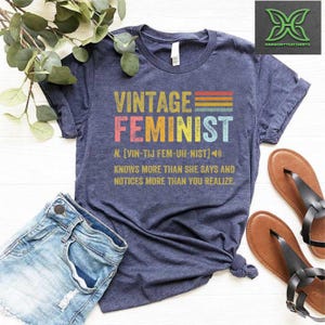 May include: A navy blue t-shirt with a vintage-style graphic that reads "Vintage Feminist" with a rainbow color scheme. The graphic also includes a definition of the word "feminist" with a phonetic pronunciation.