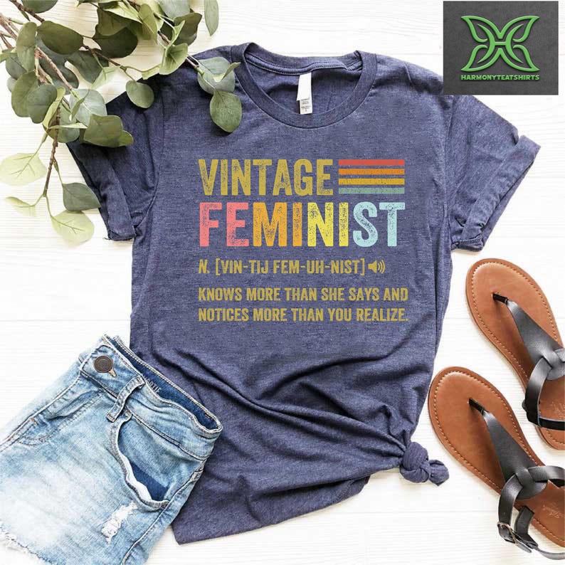 May include: A navy blue t-shirt with a vintage-style graphic that reads "Vintage Feminist" with a rainbow color scheme. The graphic also includes a definition of the word "feminist" with a phonetic pronunciation.