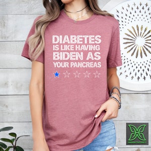Op de afbeelding: Een roze T-shirt met de tekst "Diabetes is like having Biden as your pancreas" in wit met een blauwe sterrenbeoordeling van &eacute;&eacute;n ster.