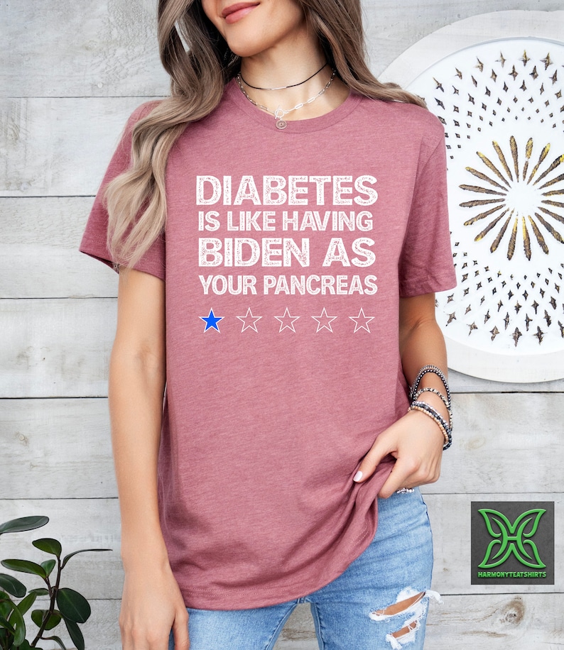 Op de afbeelding: Een roze T-shirt met de tekst "Diabetes is like having Biden as your pancreas" in wit met een blauwe sterrenbeoordeling van &eacute;&eacute;n ster.