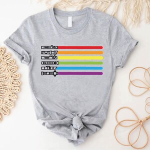 Puede incluir: Una camiseta gris con un dise&ntilde;o de arco&iacute;ris y siluetas de sables de luz negros. El arco&iacute;ris est&aacute; formado por rayas rojas, naranjas, amarillas, verdes, azules, &iacute;ndigo y violetas.